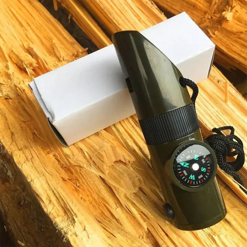 An olive green pocket compass with a black dial and black lanyard, resting on a rustic wooden surface next to its white cardboard box.